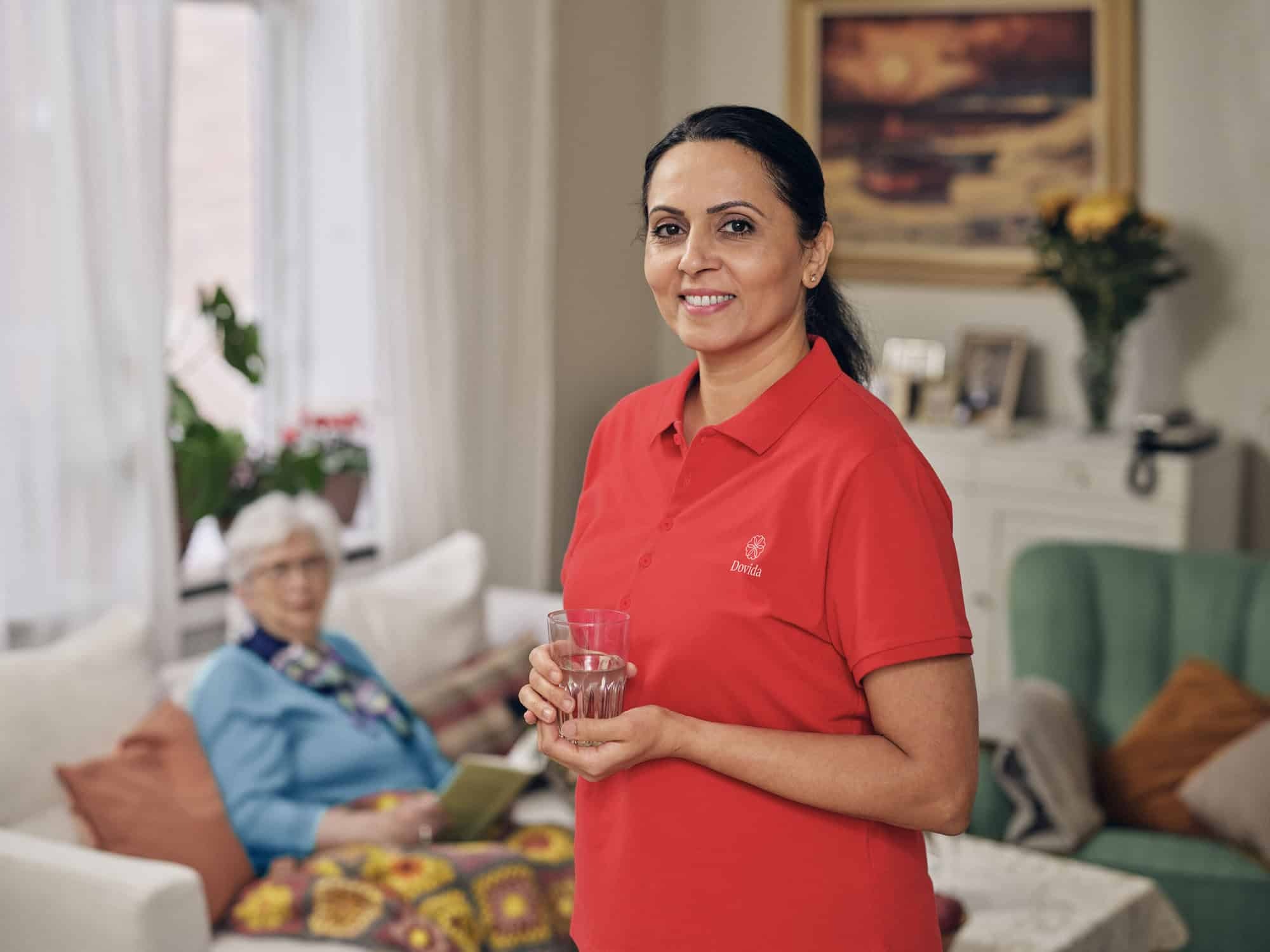 Thuiszorgverlener in rood Dovida shirt lacht terwijl ze een glas in haar hand houdt en thuiszorgdiensten vertegenwoordigt voor ouderen in Nederland. 