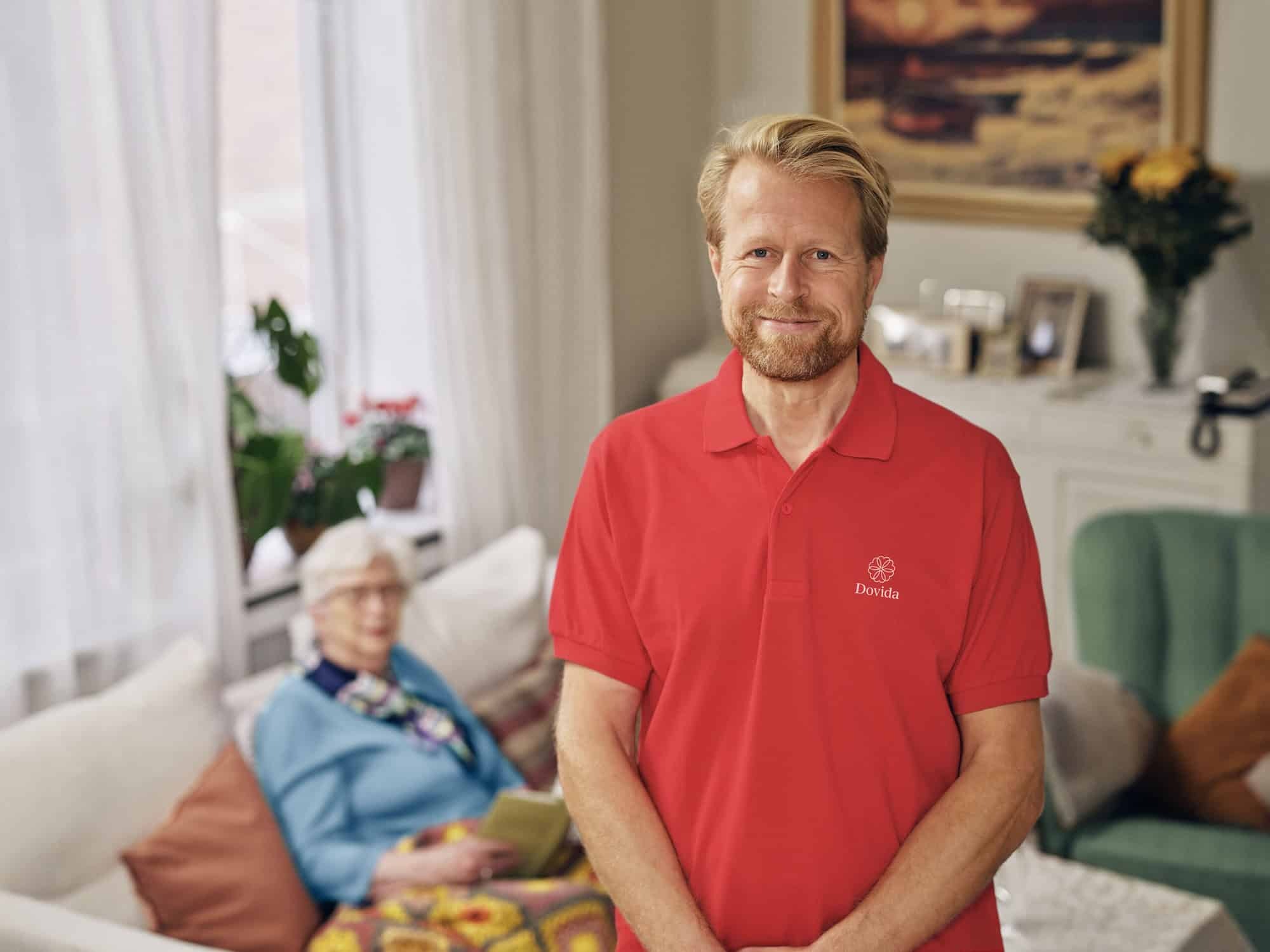 Een thuiszorg caregiver in een rood Dovida shirt staat vertrouwd, klaar om ouderen te helpen.