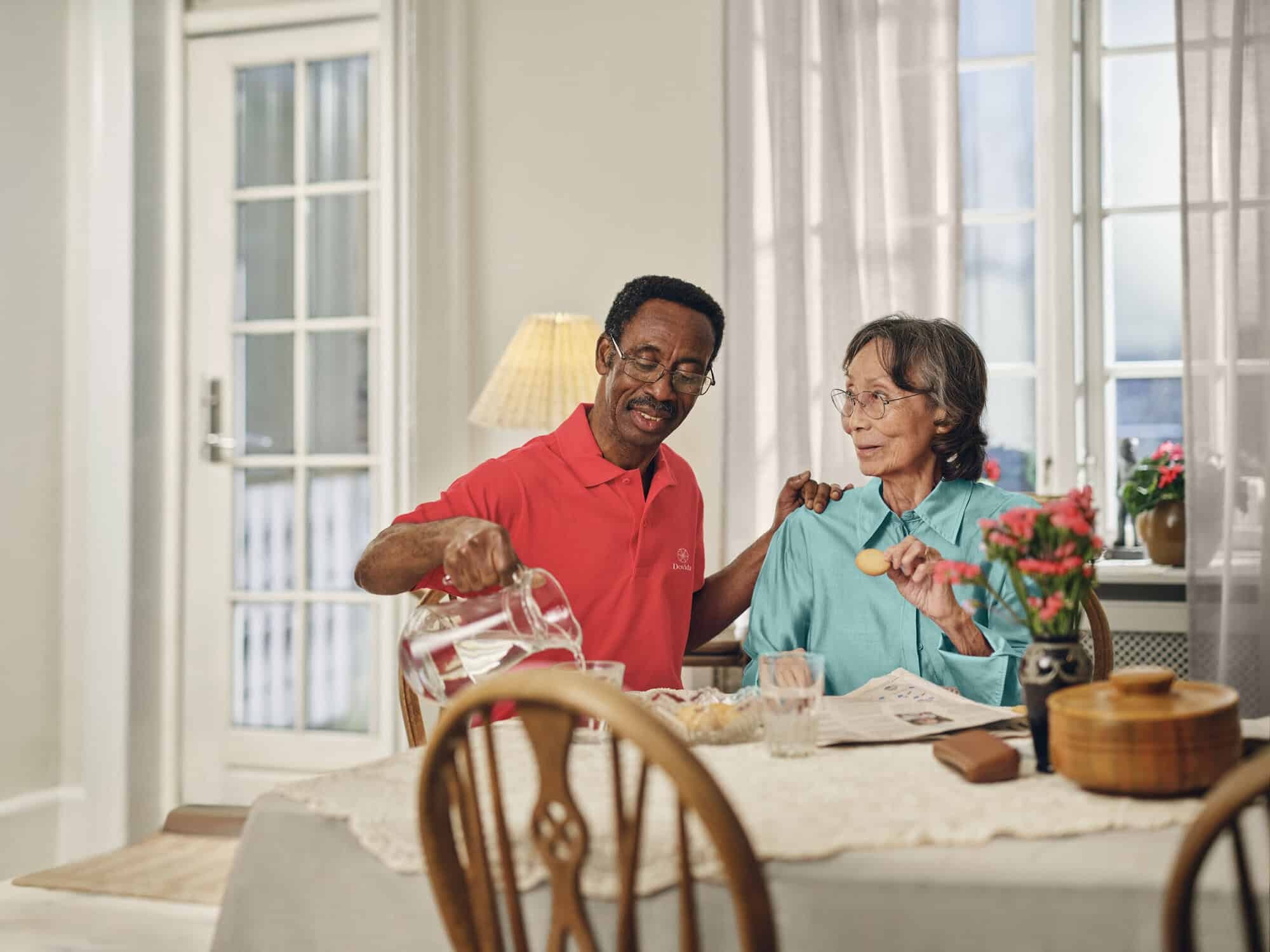 Caregiver schenkt bij de eettafel water in voor een oudere vrouw en symboliseert daarmee de persoonsgerichte thuiszorgdiensten. 